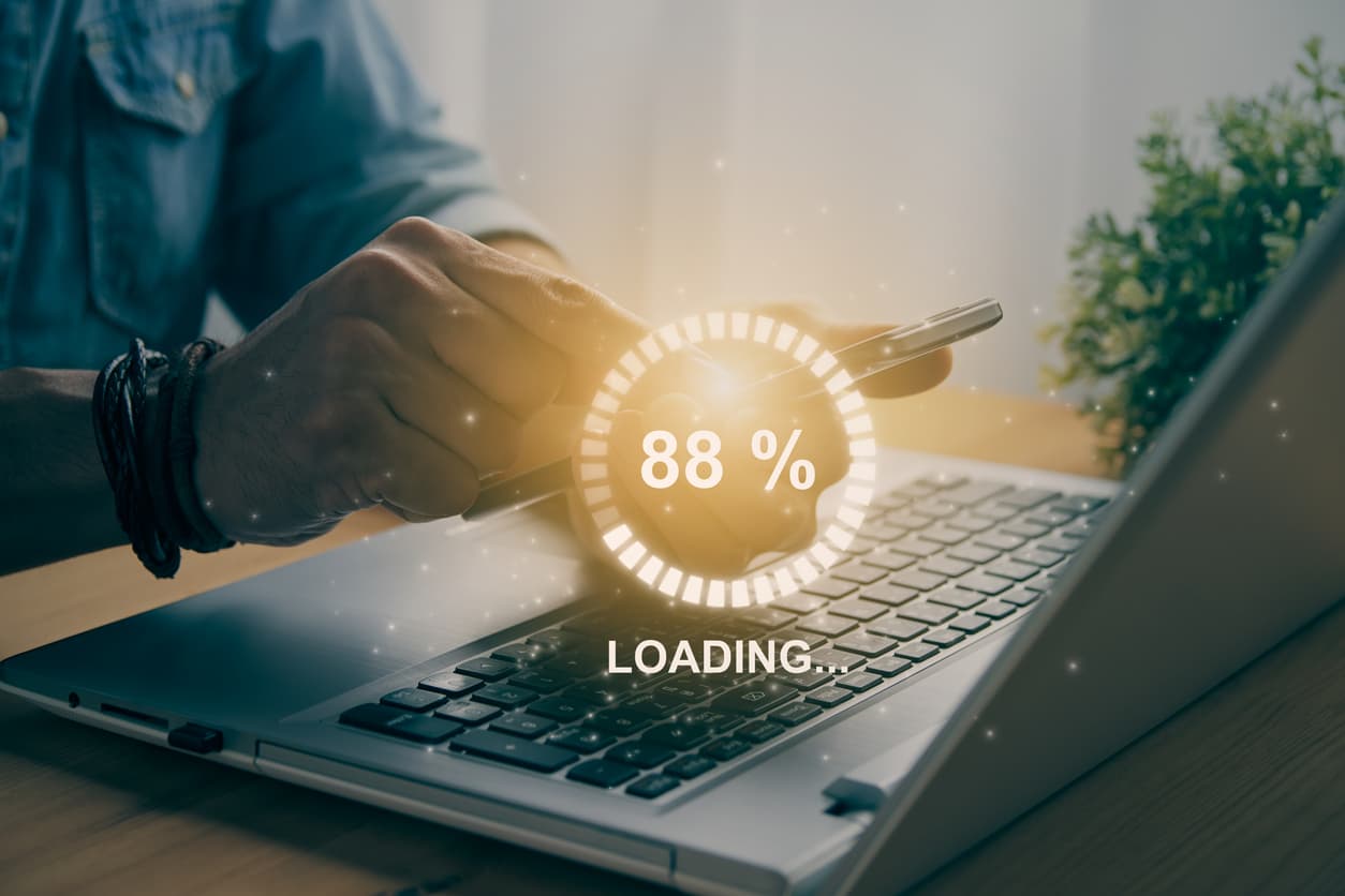 Man holding phone and working on laptop, loading circle on screen for downloading or searching.